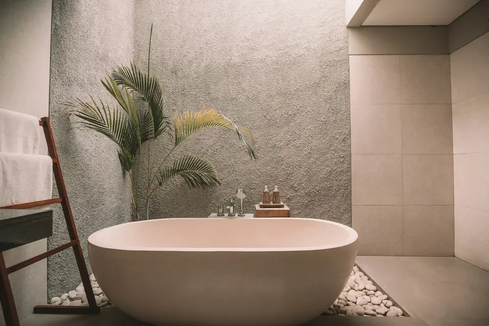 A bathroom with a freestanding tub on river pebbles, a rough plaster wall and a palm in the corner