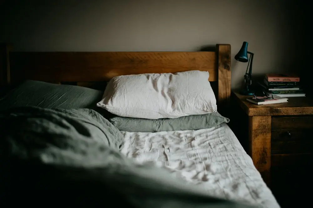 A wooden bed with sage green bedding in a dark bedroom, with a lamp and a stack of books beside it